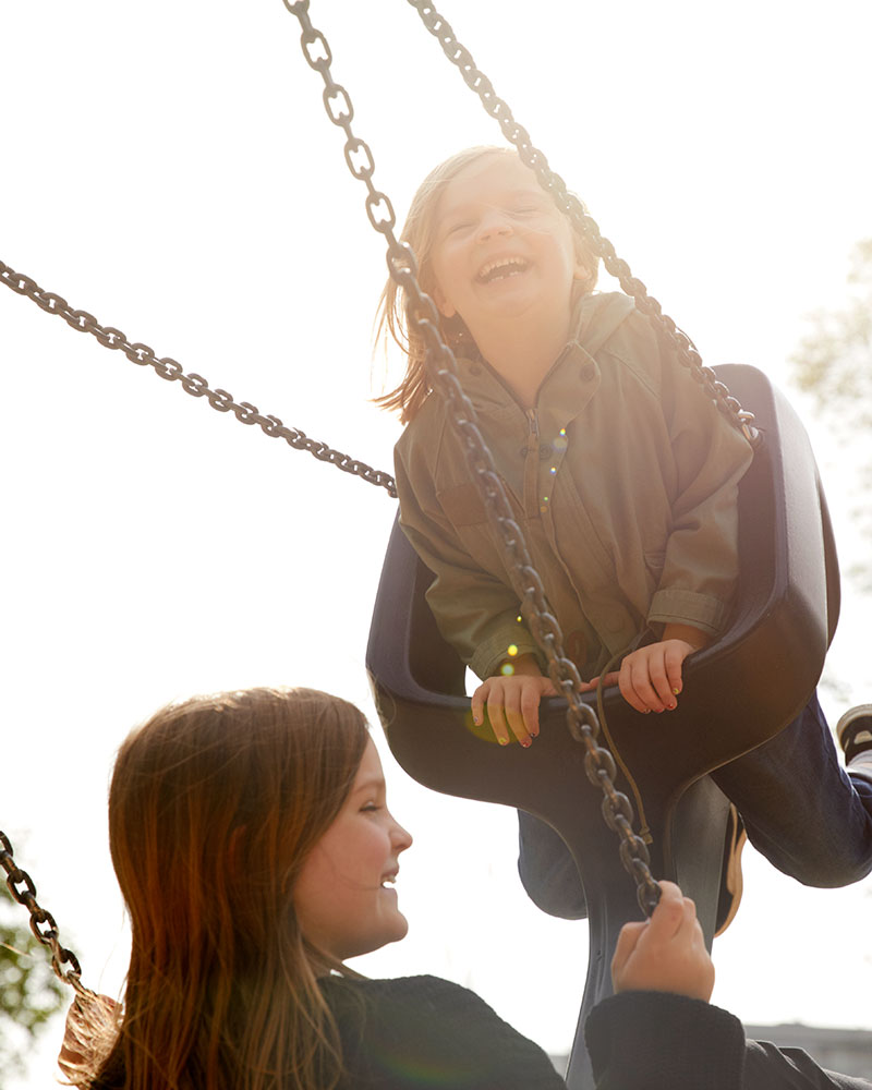 Kaksi tyttöä käyttää Tango Swing Seat -istuinta, he ovat ilmassa ja keinussa, hymyilevät ja nauttivat leikkikenttäkokemuksesta.